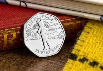 The Rupert Bear Brilliant Uncirculated 50p in display card leaning on a red and gold book spine on a wooden surface